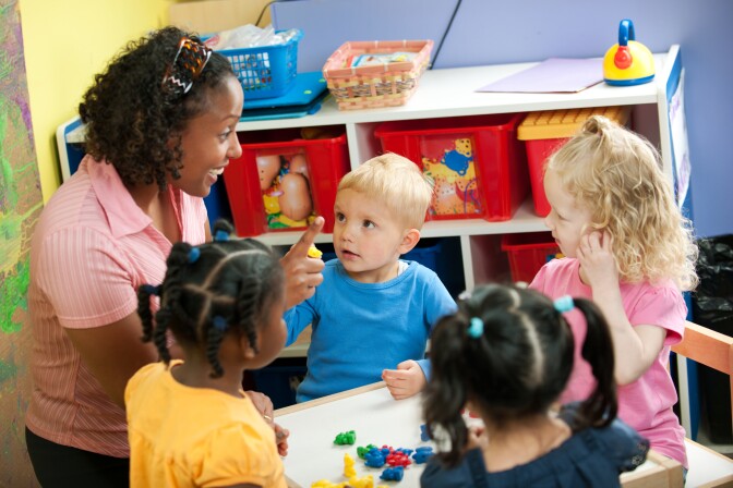 Small children at a preschool center. A teacher holds up one finger as she counts.