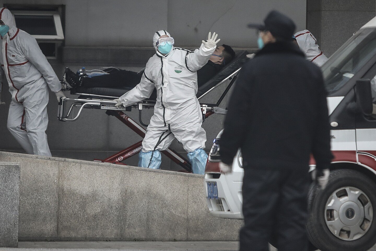 Two medical workers in white personal protective gear wave as they wheel a face mask-wearing man in a stretcher.
