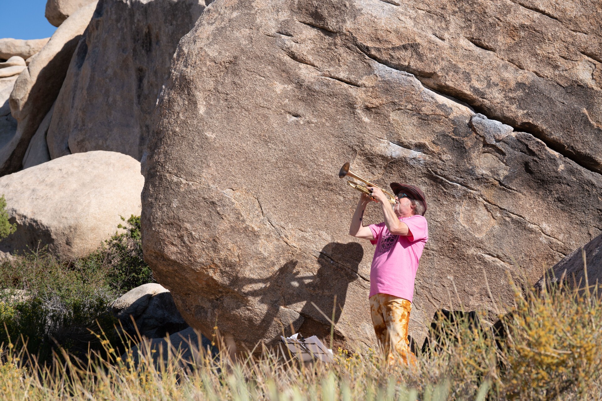 Andrew Spence is wearing a bright pink t-shirt, orange tie-dye pants and a wide-brimmed brown hat as he plays the trumpet, pointing the instrument upwards as he plays. He's playing next to a large rock formation where his shadow is cast on the rock, a mirror silhouette of him playing the trumpet. In the foreground, dry grass line the bottom of the photo.