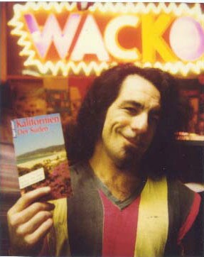 Billy Shire inside WACKO, an extension to the Soap Plant. Behind him is a bright neon sign with the word "WACKO" in vibrant letters. He's holding a brochure that reads, "Kalifornien Der Soden."