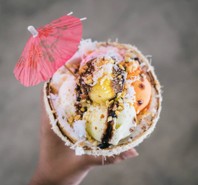 An overhead photo of a coconut ice cream sundae. The sundae is topped with a pink paper umbrella, coconut shavings and chocolate syrup drizzle.