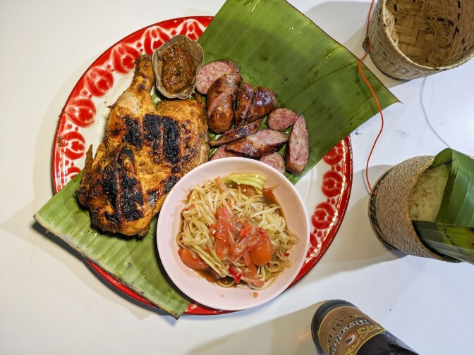 Lao sausage and chicken with papaya salad plate served with sticky rice and beer on the side. The chicken is a thigh and leg with a slight char on the skin. The Lao sausage is cut in thin slices.
