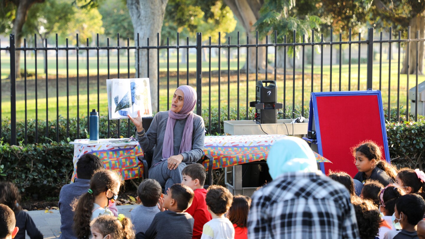 A woman wearing a headscarf holds up a picture book and reads to a group of children sat down in front of her. The reading is taking place in an outdoor area. Behind the woman and group of children is a black iron fence. Beyond that is an open area of green grass and tall trees.