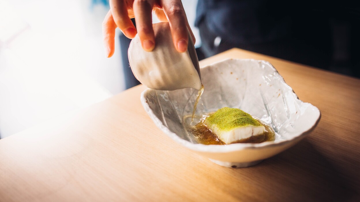 An asymmetrical ceramic dish holds a small, bite-sized piece of white steamed fish sitting in a thin, broth-y sauce. The fish is topped with a fine green powder. Additionally, someone is pouring more of the sauce from a small ceramic container.
