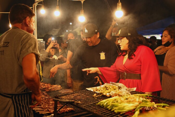 A man in an L.A. cap and black shirt and a woman wearing red point to barbecue sticks at a night market.