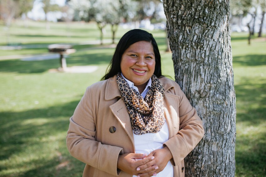 Jessica Cardenas, a Latine woman with long black hair wearing a beige coat, smiles in front of a tree.