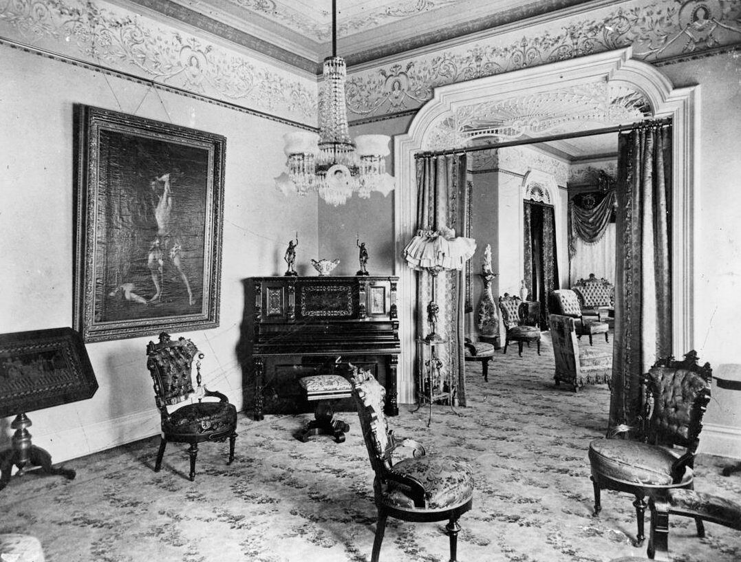 A photograph of Arcadia Bandini Stearns de Baker's front parlor. A sitting room with several ornate chairs and an upright piano is pictured, with a chandelier hanging from the ceiling above.