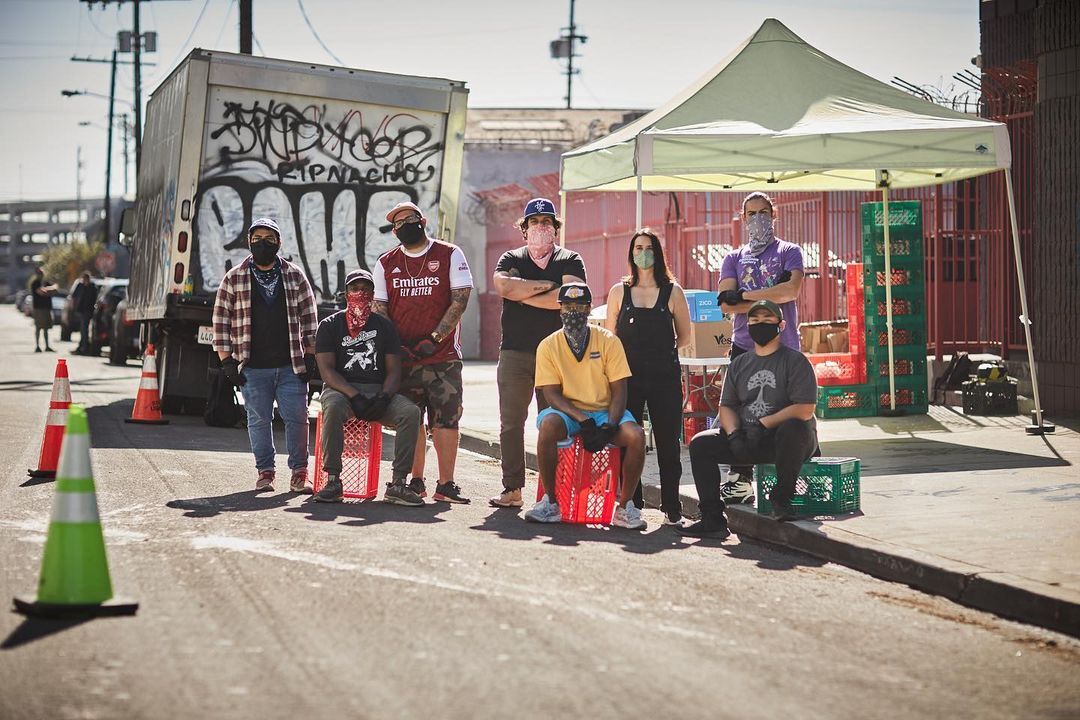 A group of volunteers at No Us Without U stand for a group photo along a curb in Los Angeles. Some of the volunteers are sitting on plastic produce crates while others stand behind them with their arms crossed or at their side. Behind the group of volunteers is a collapsable outdoor tent where there are stacks of plastic produce crates and a plastic table for distributing food. Parked along the curb is a small moving truck with graffiti on the back.