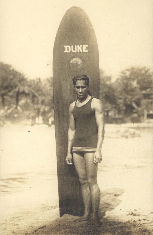 A sepia-toned old photo of a young Duke Kahanamoku standing in front of a wood surfboard, propped up on the sand. The surfboard says, "DUKE" across the top. Duke Kahanamoku is wearing a men's bathing suit and is looking at the camera.